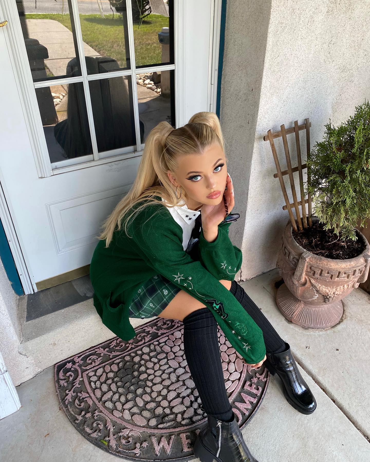 a young woman, loren gray, is sitting on a doorstep. she has blonde hair styled in pigtails and is wearing a green sweater, a plaid skirt, kneehigh socks, and black boots. she is looking at the camera. behind her is a white door and to her right is a potted plant. she is sitting on a doormat that reads "welcome."