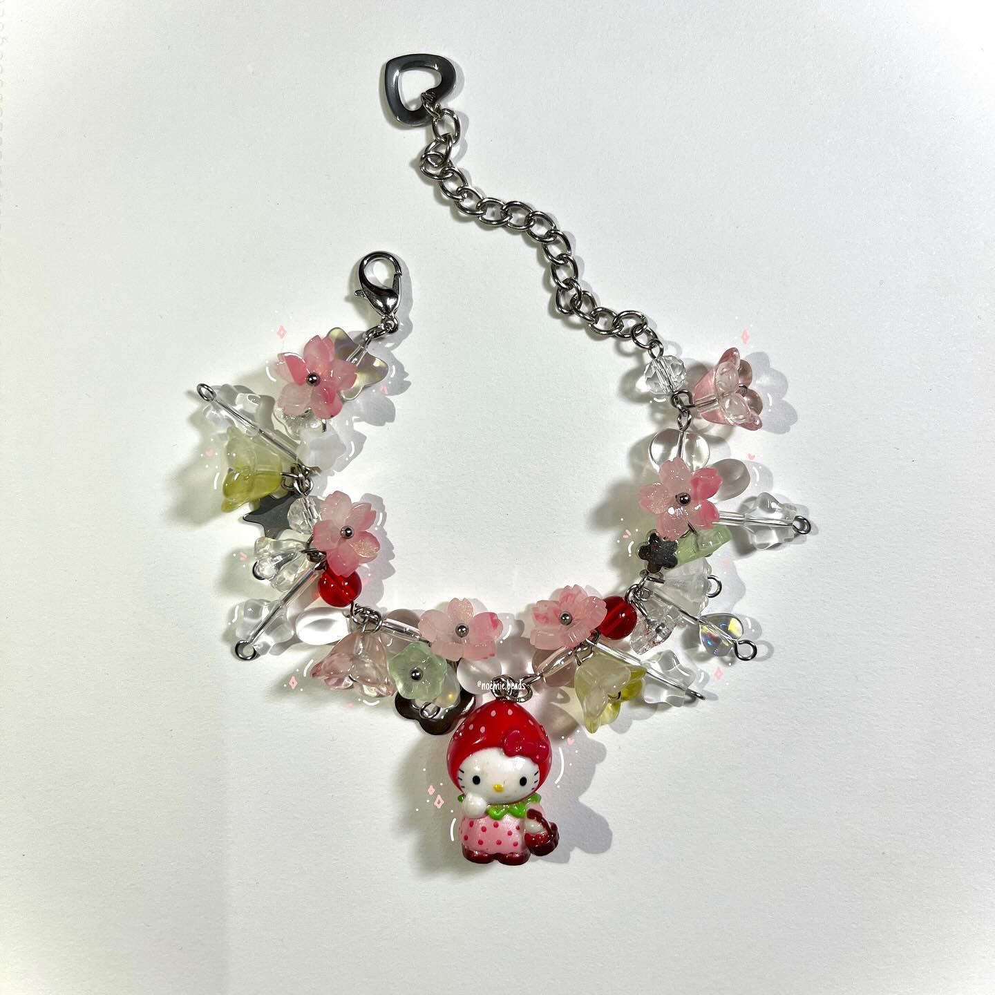 a charm bracelet featuring pink flower beads, red and clear beads, and a hello kitty charm dressed as a strawberry, lying on a white surface. the bracelet has a silver chain and a clasp.