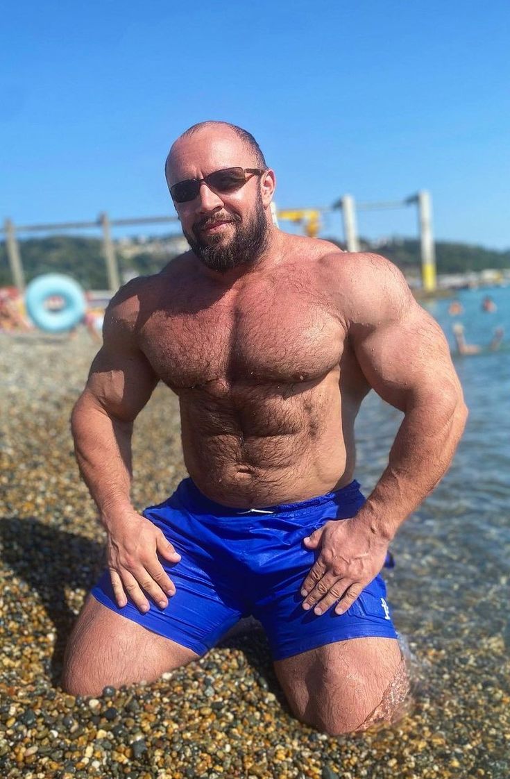 a muscular, hairychested bald man wearing sunglasses and blue swim shorts kneels on a pebble beach near the ocean. he appears to be enjoying a sunny summer day. his physique suggests he is a bodybuilder or strength athlete. the overall mood is one of relaxation and enjoyment of the outdoors.
