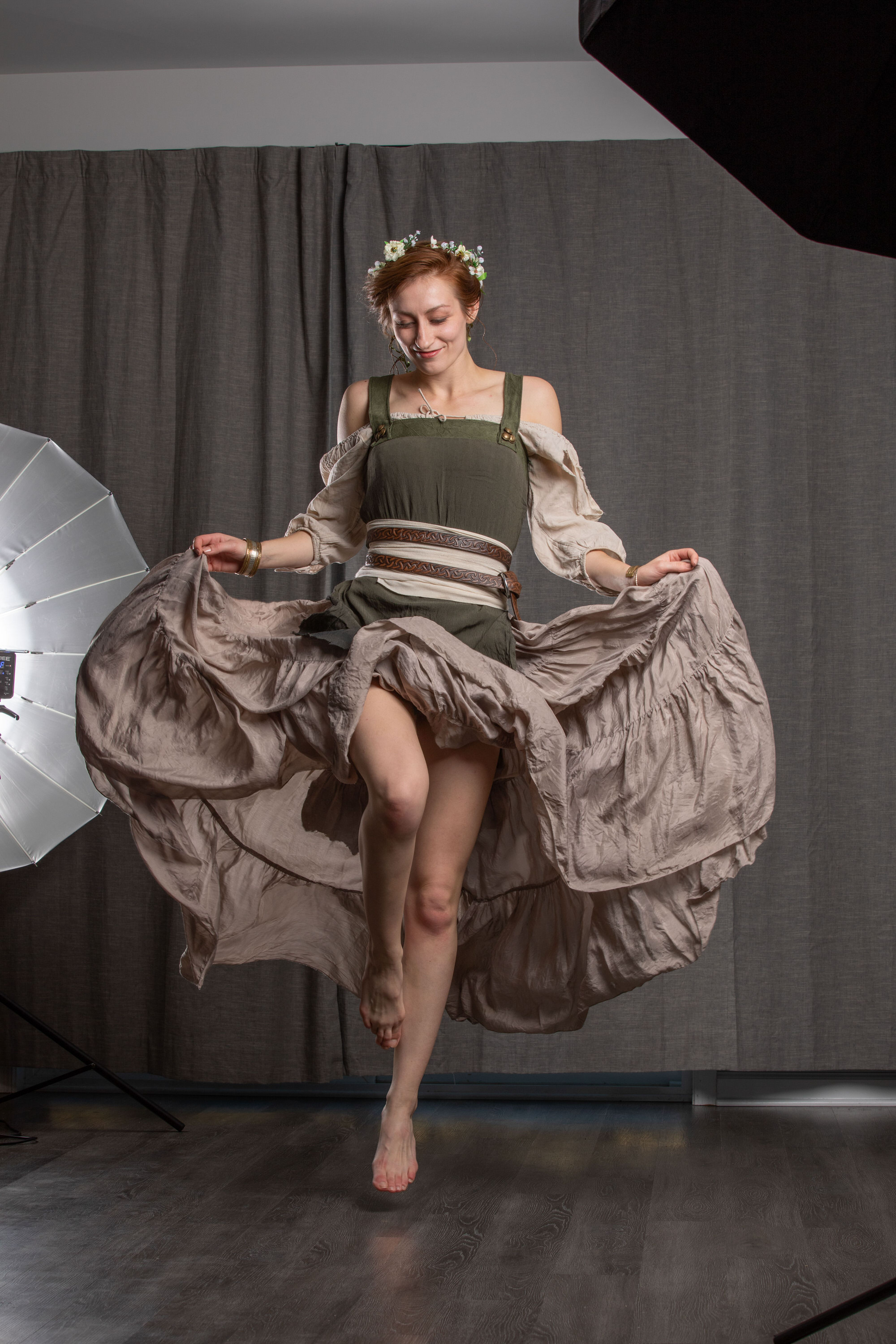 a woman in a flowing dress jumps in a photography studio. she is illuminated by a softbox to her left, casting a soft light on her. the background is a dark grey curtain, and the floor is a medium brown wood. she is wearing a green bodice with white sleeves and a brown belt, and her skirt is a layered, flowing light brown fabric. she has a flower crown on her head and is smiling. the overall feeling of the image is one of joy and movement.