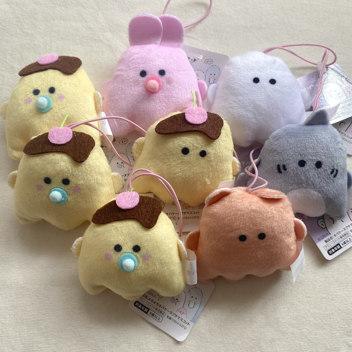 the image shows a collection of small, plush toys, each with a string loop for hanging. there are three yellow puddingshaped characters with brown "pudding" tops and colorful pacifiers. there's also a pink bunny with large ears and a pacifier, a white ghostlike figure with simple black eyes, a gray shark with stitched gills, and an orange creature with small ears. each toy has a paper tag attached.