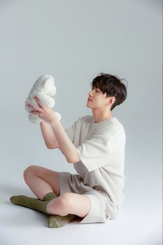 a young asian man with short black hair is sitting on the floor and holding a white teddy bear. he is wearing a white tshirt and shorts and green socks. the background is a solid white.