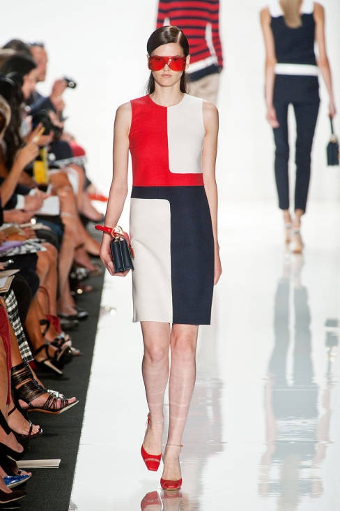 a model walks the runway in a red, white, and blue colorblocked dress and red sunglasses. she is carrying a black handbag. the audience is seated on the left side of the runway, and another model is walking in the background.
