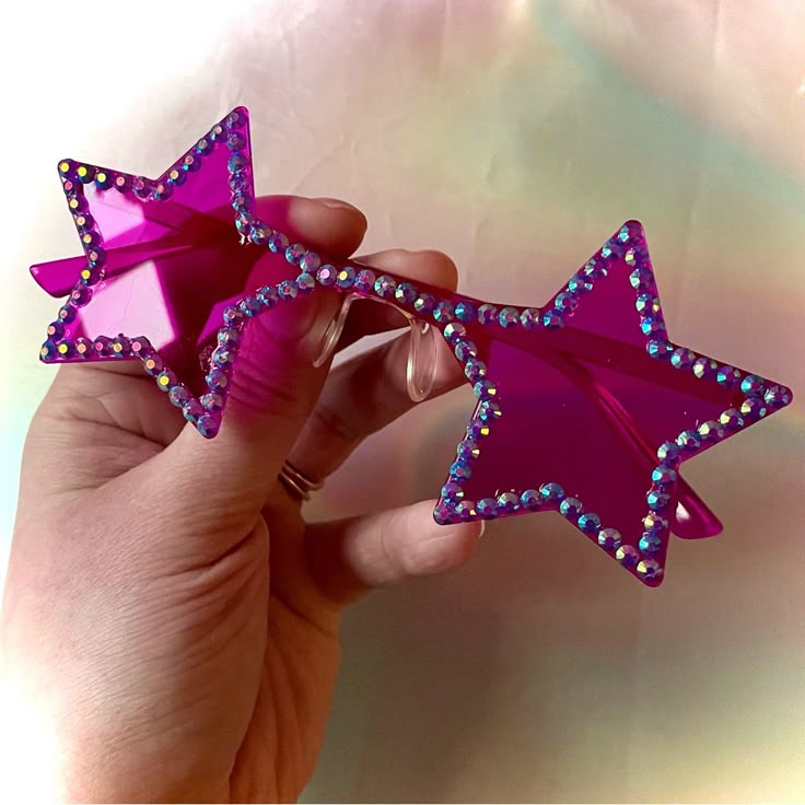 a hand is holding a pair of pink, starshaped sunglasses. the edges of the star shapes are lined with small, shiny jewels.