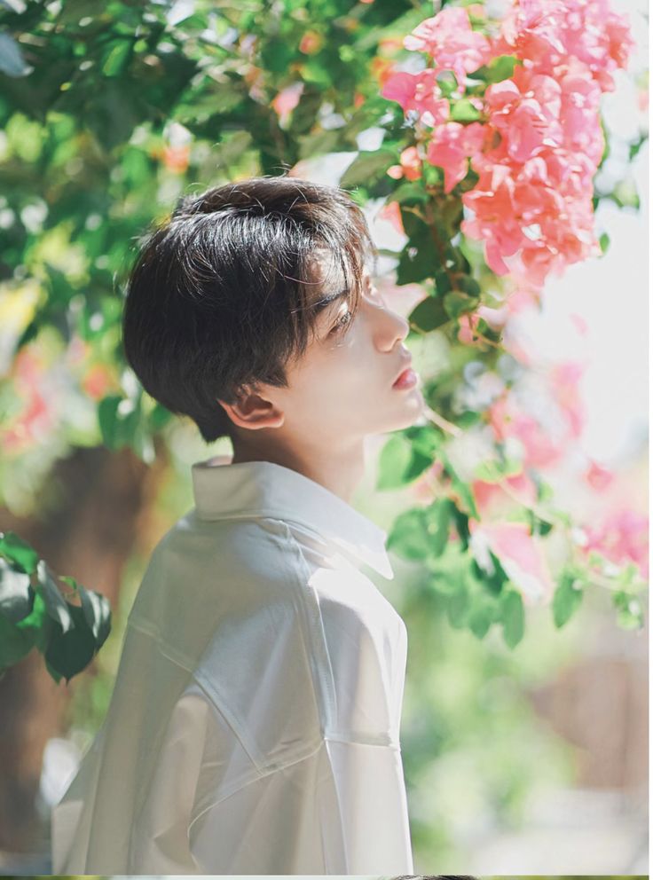 a young man with short black hair wearing a white shirt is captured in a serene outdoor setting. he stands amidst vibrant green foliage and delicate pink bougainvillea flowers, his face tilted upwards towards the sunlight filtering through the leaves. the image evokes a sense of tranquility and peacefulness.