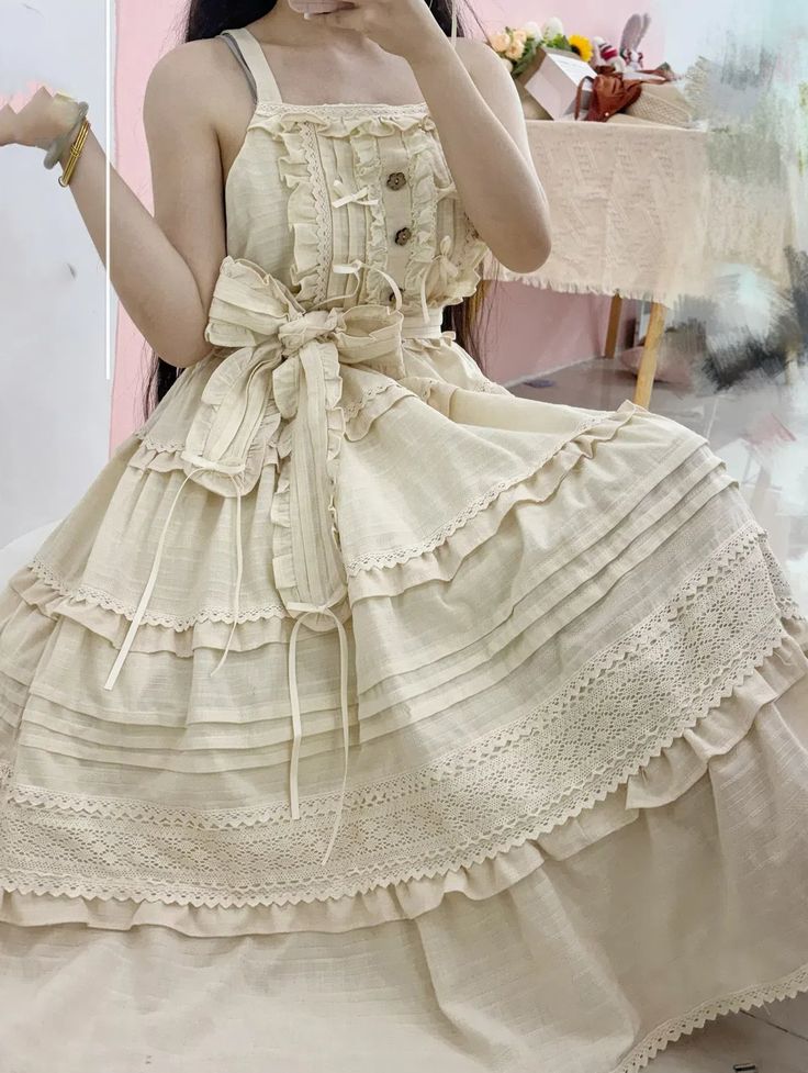 a young woman poses for a mirror selfie, wearing a long, flowing beige dress with intricate lace and ruffle details. the dress features a square neckline, delicate buttons, and a large bow at the waist. the romantic and feminine style of the dress evokes a vintage, cottagecore aesthetic. the photo is taken indoors in a naturally lit room, likely a bedroom, and the perspective is at eye level.