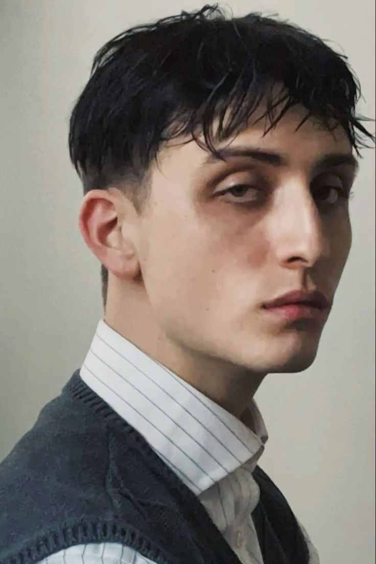 the image is a closeup portrait of a young man with dark, wetlook hair styled short and slightly textured. he's wearing a crisp white pinstripe shirt with the collar neatly buttoned and a dark grey sweater vest. his expression is serious and somewhat pensive. the background is a plain, light grey, drawing focus solely to the subject. the overall mood is one of stylish simplicity.