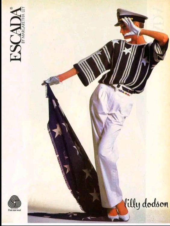 a vintage advertisement for escada clothing features model lilly dodson. she wears a navy blue and white striped sweater with star accents, white highwaisted pants, white gloves, and a navy blue captain's hat. she poses dramatically, holding a long piece of blue fabric with white stars printed on it. the background is plain and offwhite.