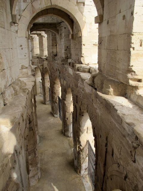 the image shows a highangle, closeup view of the interior passageways of the arènes de nîmes, a roman amphitheater in nîmes, france. the scene is dominated by a long, narrow corridor lined with numerous arches and decaying stone walls. the arches create a sense of depth, leading the eye deeper into the structure.  the stone is light beige in color and shows significant weathering and age, indicating the structure's antiquity.  light and shadow play across the walls, highlighting the texture and details of the stonework. a black metal gate is partially visible near the bottom, partially obscuring a view to another section. the overall impression is one of historical grandeur and the passage of time.