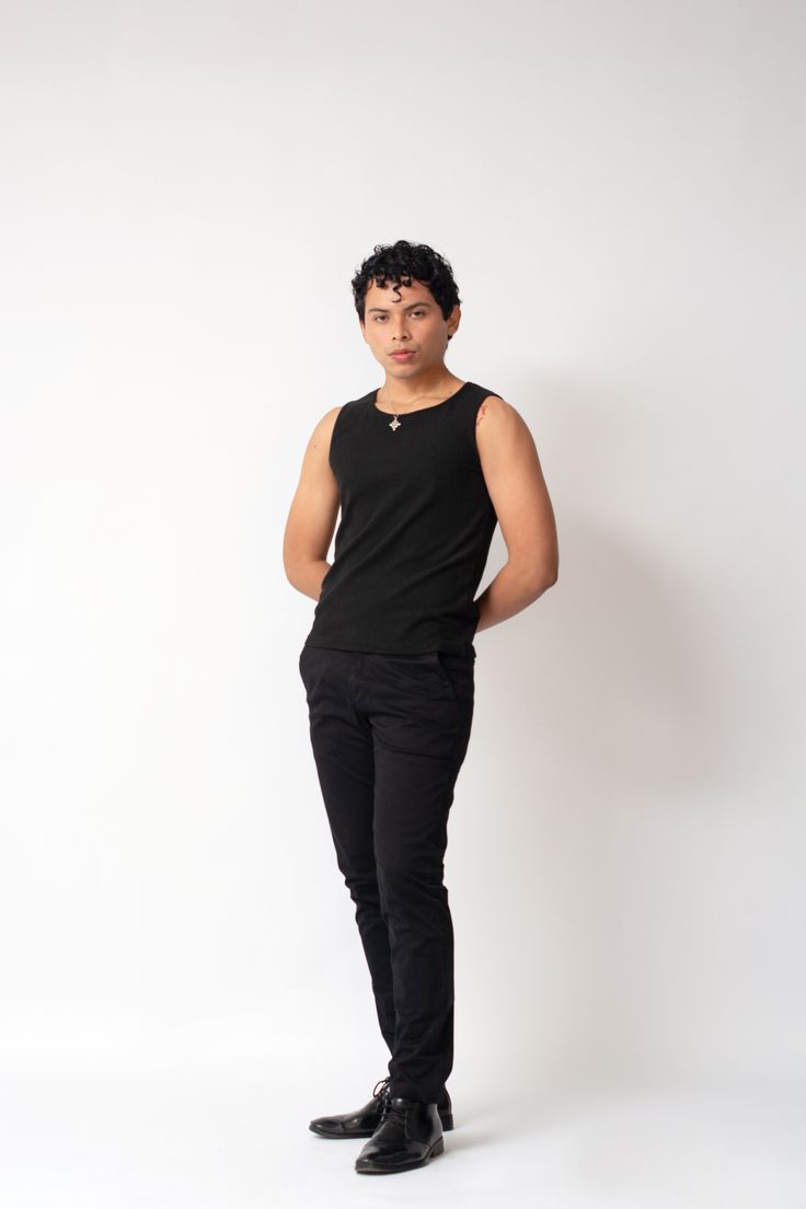 a fullbody studio shot of a young hispanic man standing against a white background. he is wearing a black tank top, black pants, and black dress shoes. his hands are in his pockets. he has a neutral expression on his face.