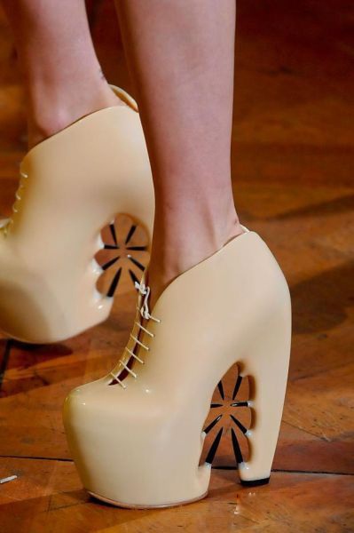 the image shows a closeup of a person wearing a pair of avantgarde, beige platform high heels. the shoes feature a unique design with laces in the front and decorative cutouts on the sides, exposing parts of the foot and the background. they are shiny and appear to be made of a smooth material. the shoes are set against a wooden floor.