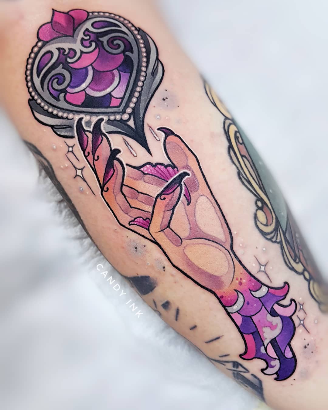 a closeup image of a neotraditional tattoo on someone's arm. the tattoo depicts a hand with purple scales and long, pointed black nails. the hand reaches up to grasp an ornate heartshaped locket encrusted with jewels. the locket is colored in shades of purple and pink with white highlights. small white dots surround the hand and locket, giving the impression of glitter or magical sparkle. the tattoo is done in a bold, graphic style with black outlines and shading. the artist's signature, "candy ink", is visible in white ink on the arm below the tattoo.