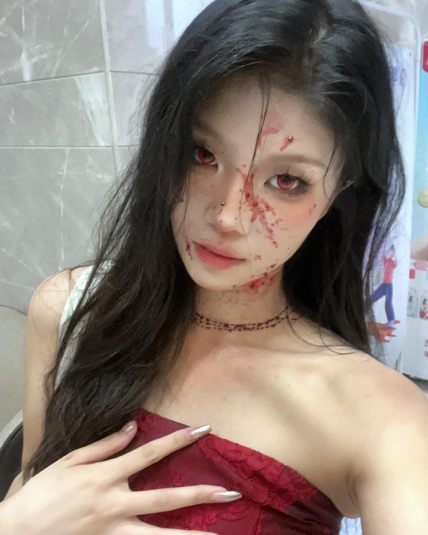a young woman takes a selfie in the mirror showcasing her halloween costume and makeup. she is dressed in a red lacy top and has fake blood splattered across her face and chest. her makeup includes red contact lenses, further enhancing the spooky vampirelike look she embodies.