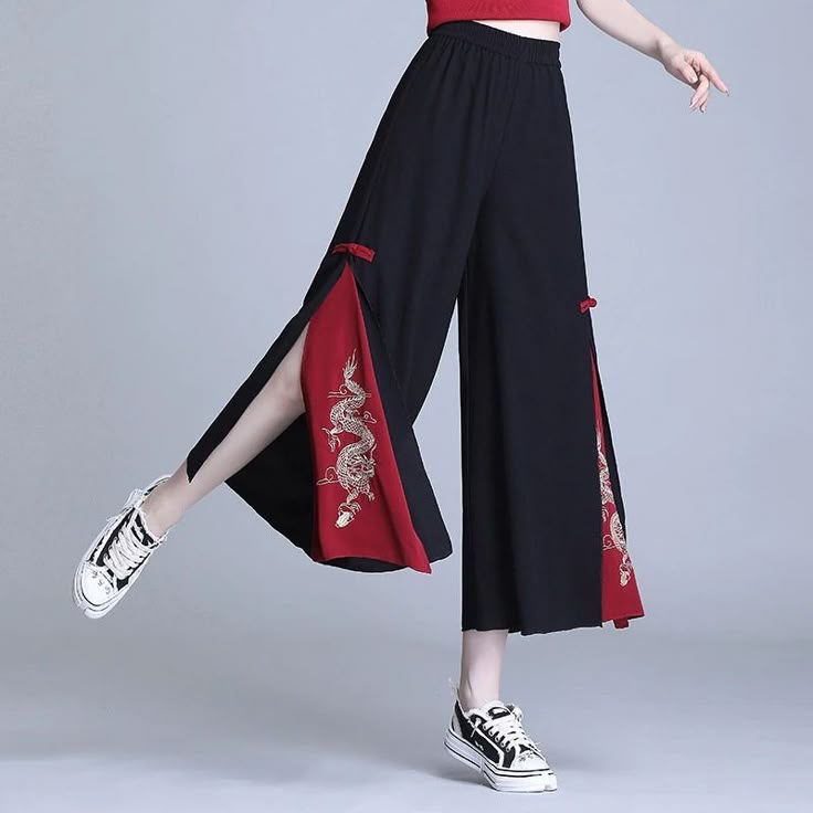 a person wearing black wideleg pants with red trim and embroidered dragons on the sides. they are paired with white sneakers. the person is standing on a gray surface.