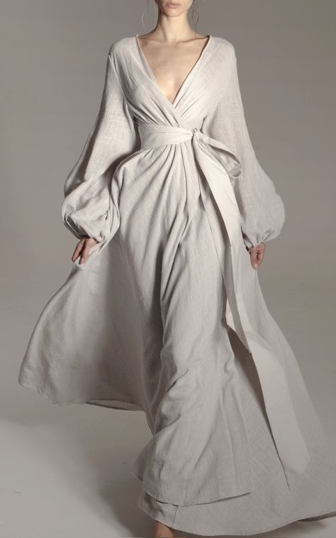 a woman is standing, wearing a flowing, fulllength, light grey linen dress with a deep vneckline and long, billowing sleeves. the dress is cinched at the waist with a matching tie belt. she is posing against a light grey backdrop in a photography studio setting. the lighting is soft and diffused, highlighting the flowing silhouette of the dress and the natural texture of the linen fabric.