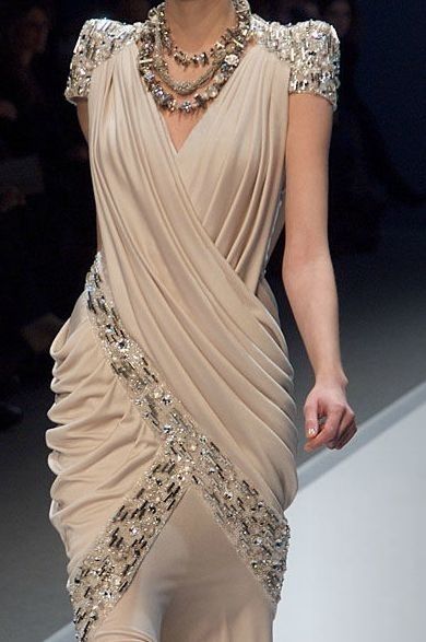 a model is walking down a runway, wearing a creamcolored gown with sequins. the gown is designed in a grecian style, featuring a draped bodice and a flowing skirt. the model is also wearing a thick silver necklace. she is looking at the camera with a confident expression. the background is out of focus, showing other models and members of the audience. the overall image is one of elegance and sophistication.