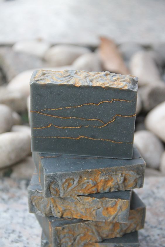a stack of four gray soap bars with gold flecks on a rocky background. the soap bars are arranged vertically, with the largest bar at the top and the smallest bar at the bottom. the soap bars are made from a combination of gray and gold materials, creating a visually appealing contrast. the rough texture of the soap bars suggests that they are made from natural ingredients. the rocky background adds a touch of rustic charm to the image, suggesting that the soap bars are handmade. the image is a beautiful representation of the natural beauty of handmade soap.