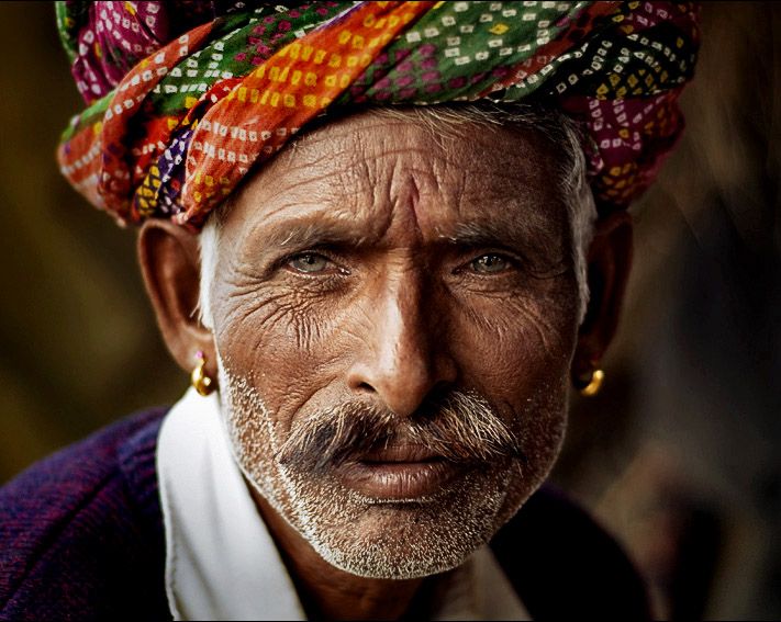 a closeup portrait of an elderly indian man wearing a colorful turban. he has a mustache, wrinkles, and gold earrings. the image highlights the details of his weathered face and captures the essence of indian culture.