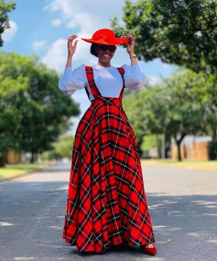 a woman is standing on a street wearing a white blouse, a red plaid skirt with suspenders, and a red hat. she is also wearing sunglasses and has her hands on her hat. there are trees in the background.