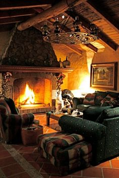 the image depicts a warm and inviting living room interior. a large stone fireplace is the focal point, with a fire burning brightly inside. an armchair and a couch provide seating, along with an ottoman. a coffee table is placed in front of the couch. a picture frame hangs on the wall. a chandelier illuminates the space. the room features wood beams on the ceiling and a stone wall. the floor is covered in tile. the warm lighting creates a cozy atmosphere.