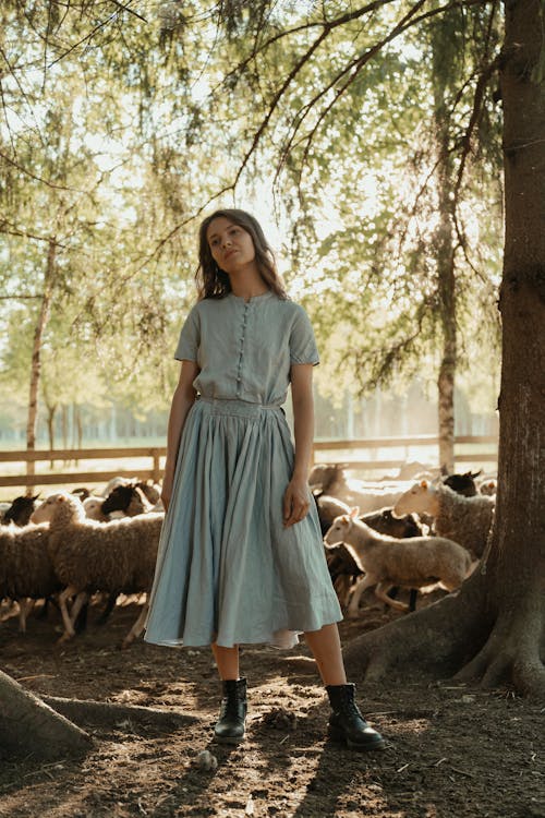 a young woman stands in a sundappled forest setting amongst a flock of sheep. she is wearing a light blue linen midi dress with a slightly gathered waistline and short sleeves, buttoned down the front. her black ankle boots are clearly visible and contrast with the dress. the dress has a somewhat vintage or rustic style. she has shoulderlength brown hair and appears calm and serene. the overall atmosphere is peaceful and pastoral.  the background includes a large tree trunk at the right edge and sunlight filtering through trees.