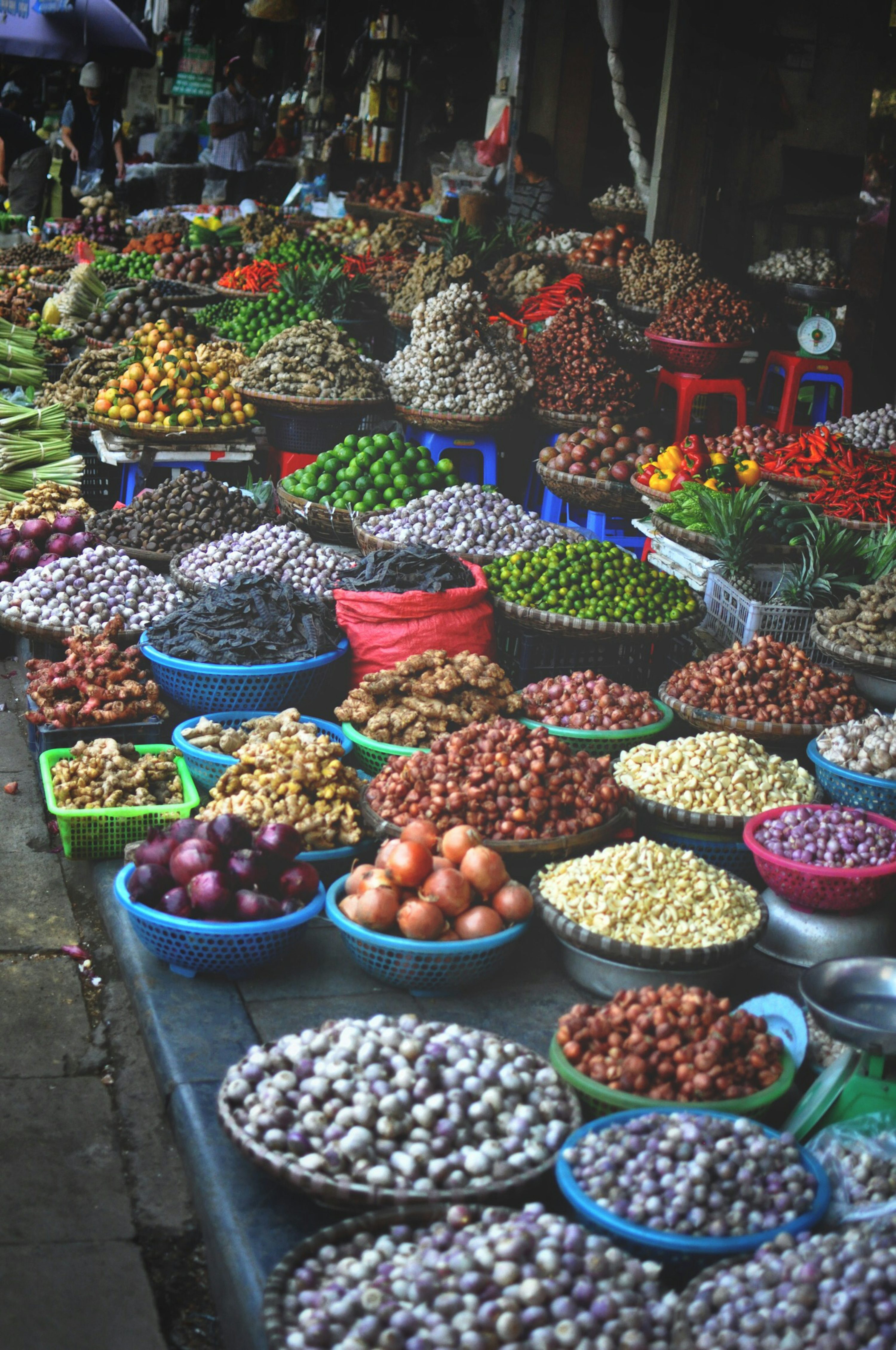 the image shows a bustling asian market overflowing with an abundance of fresh produce, spices, and other ingredients.  numerous baskets and containers are piled high with a diverse array of items including garlic, ginger, onions, various beans, peanuts, chilies, limes, and other herbs and vegetables. the scene is vibrant and colorful, showcasing the rich textures and variety of food available. the market setting suggests a busy street market in southeast asia, likely vietnam, based on the types of produce displayed. the overall impression is one of abundance, freshness, and the bustling energy of a traditional market.