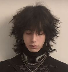 this is a portrait of a young man with long black hair styled with a fringe, wearing a black choker with several chains hanging from it, and two silver earrings. he has closed his eyes and is looking down thoughtfully. his style is cool and stylish, with a dark and edgy vibe.