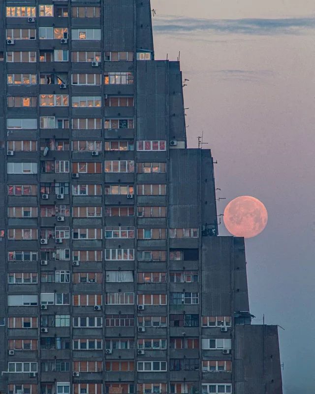 a closeup shot of a tall, grey apartment building with numerous windows. the building's facade is detailed and shows a pattern of repetitive windows.  a large, full pink moon is visible in the sky to the right of the building, partially obscured by the building's edge. the sky is a pale, pinkish hue, suggesting it's either sunrise or sunset. the overall feeling is one of serene urban beauty.