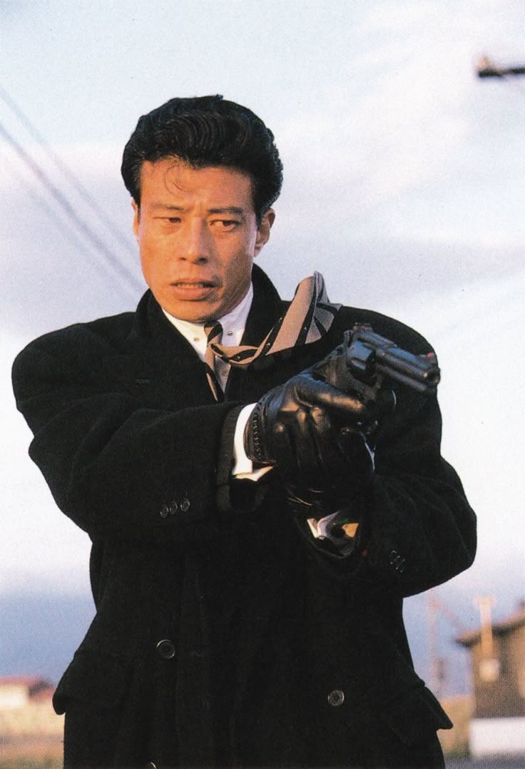 a man in a suit and overcoat holds a revolver, facing forward with a tense expression. he's wearing leather gloves. the background is somewhat blurred, showing what appears to be an outdoor urban setting.