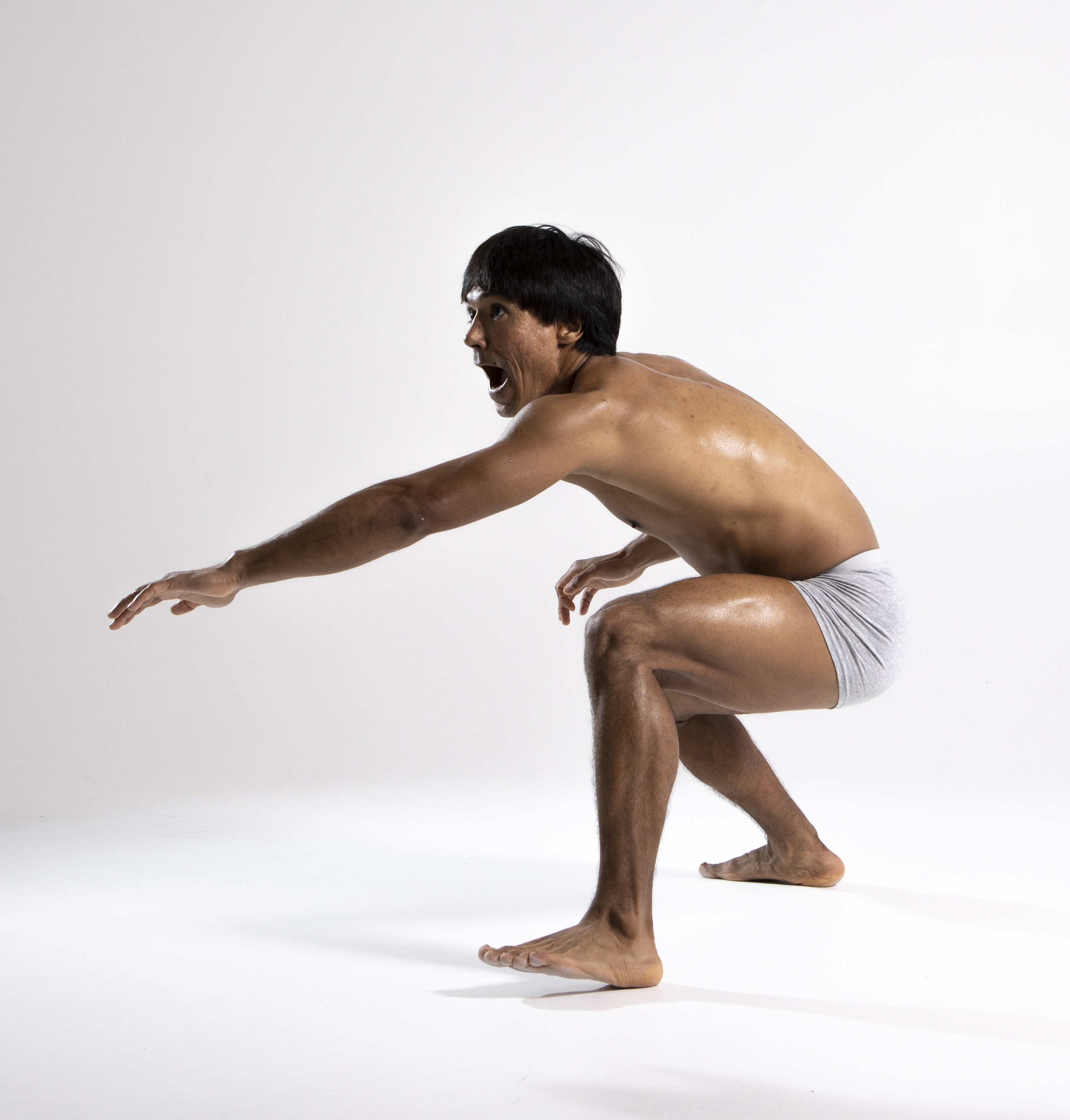 a muscular man in gray underwear is squatting with his mouth open, reaching out with his left arm. he appears to be in a studio setting with a white background.