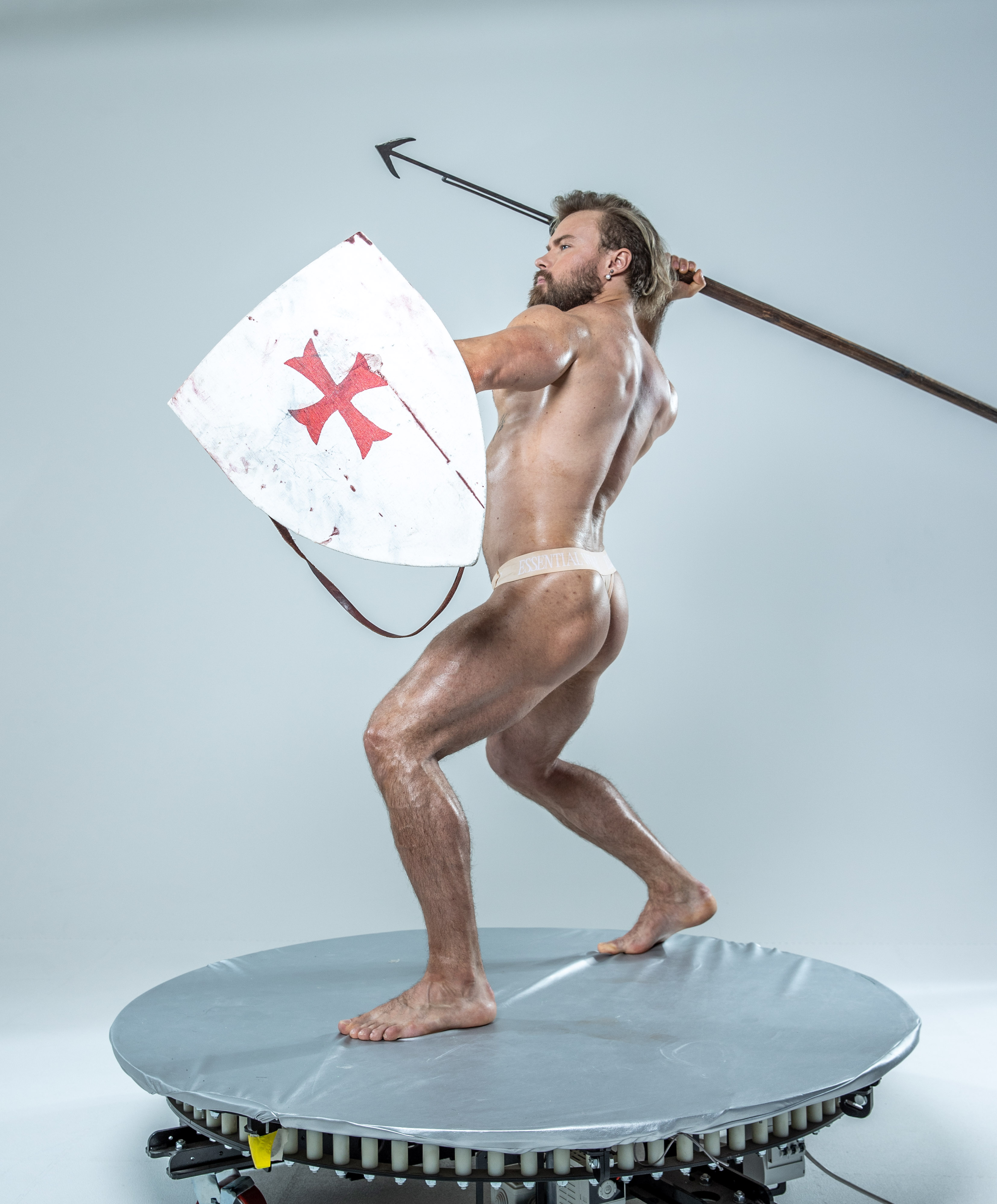 a muscular man with a beard and long blonde hair, styled in a warriorlike pose. he stands on a circular platform, wielding a spear in his right hand and a shield in his left, as if ready for battle. he is barechested, wearing only a loincloth. the image is set in a studio with plain white background, illuminated by professional lighting.
