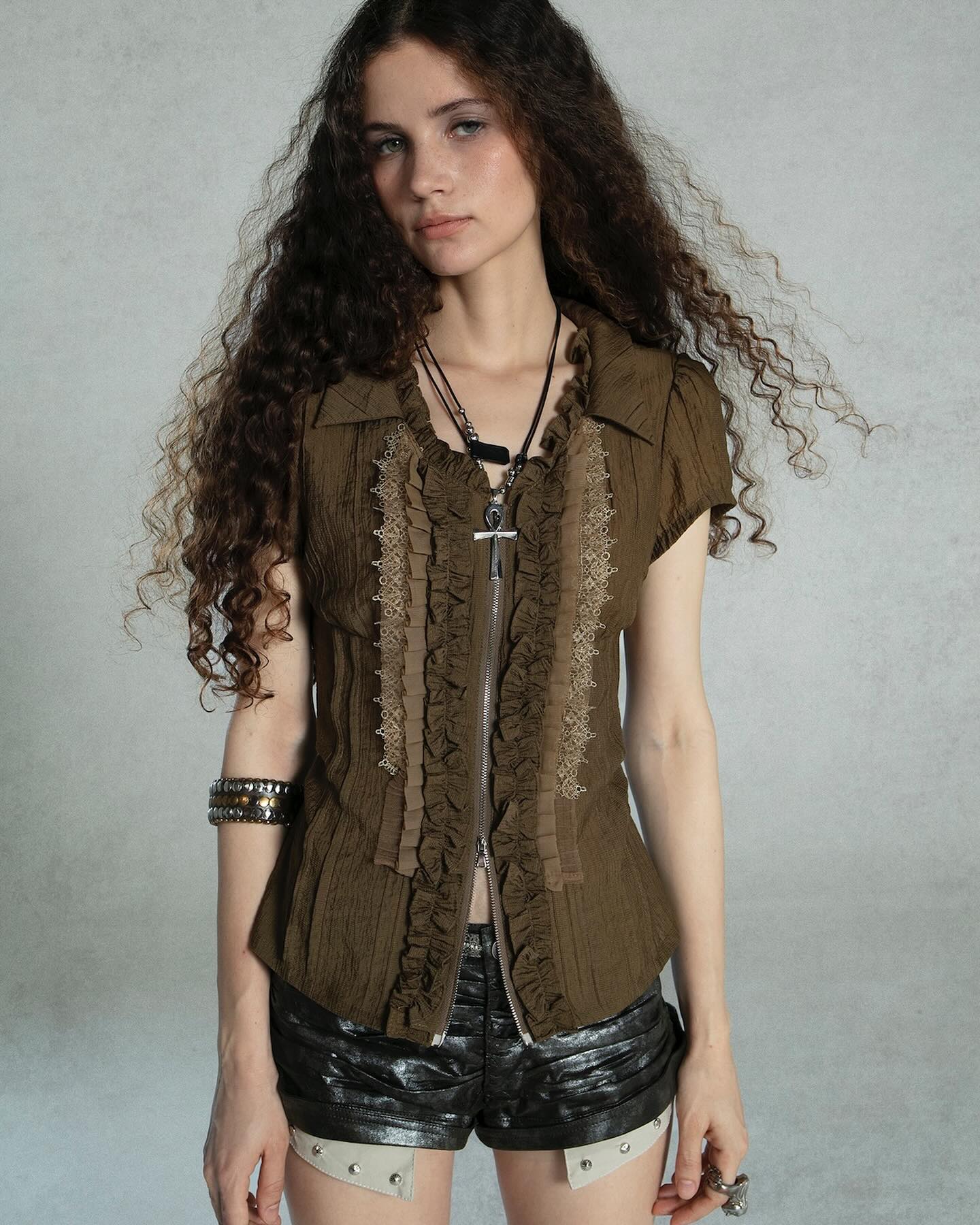 a young woman with long, curly brown hair is posing against a grey background. she is wearing a shortsleeved olive green blouse with ruffles and a zipper down the front. the blouse is layered with beige lace trim.  she's also wearing black leather shorts with studded details and several pieces of jewelry including a necklace with a cross pendant, bracelets, and a ring.  the overall style is somewhat gothic or steampunkinspired. she has a serious, slightly pensive expression.