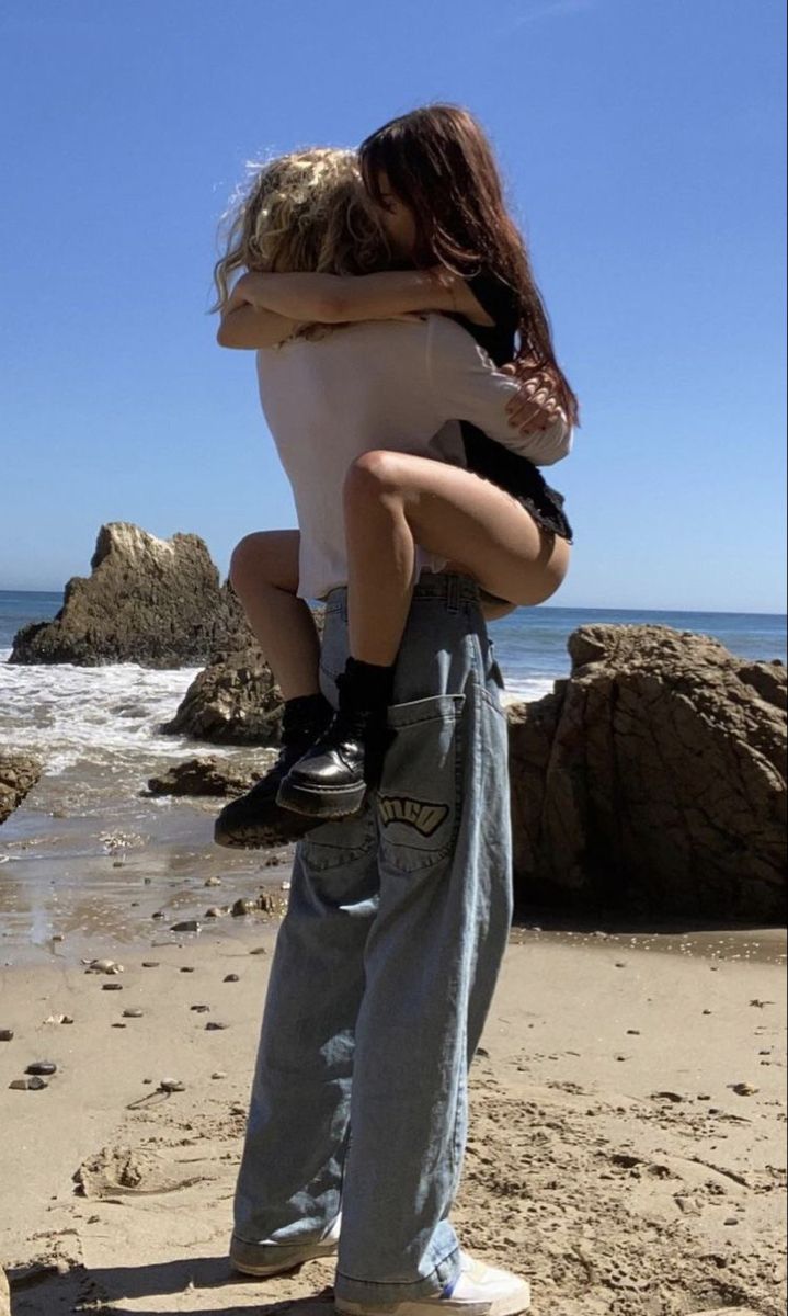 a couple is hugging and kissing on a beach. the woman is wearing a white shirt and denim jeans while the man is wearing denim jeans. the beach is sandy with rocks and water in the background. the sky is blue and the sun is setting. the couple is enjoying a romantic moment on the beach.