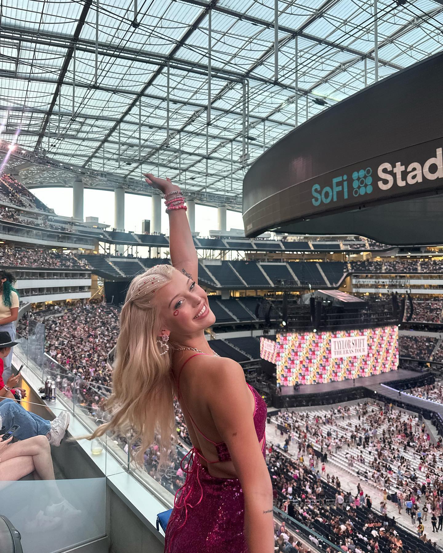 a young blonde woman in a sparkly pink openback dress is standing at a stadium concert with a crowd of people. she is smiling and waving her hand. the sofi stadium sign is visible above, and the stage for the taylor swift eras tour is visible in the background. the venue is packed with fans.