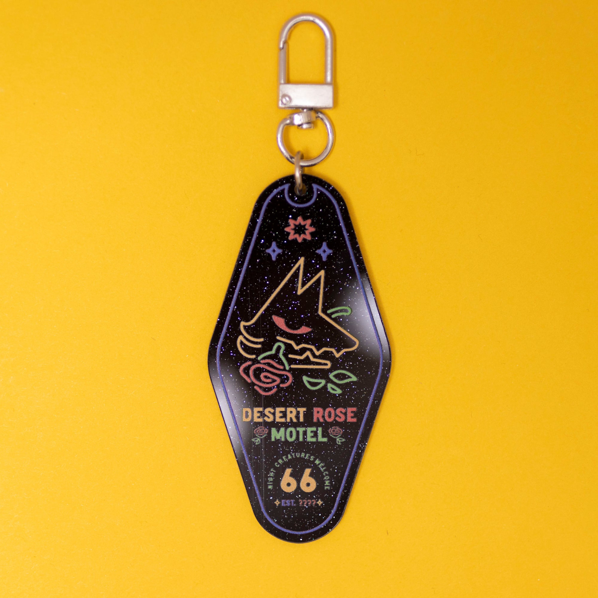 loseup shot of a motel keyshaped keychain against a solid yellow background. the keychain is black with a glitter finish, featuring a neoncolored illustration of a stylized wolf’s head with a rose, accompanied by the text "desert rose motel" and "route 66" branding. the keychain has a silver metal ring for attachment.  the overall style is retro and vintageinspired.