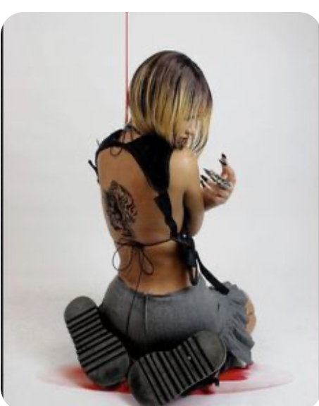 a young woman with a blonde bob haircut is photographed from behind. she is seated on a white floor, knees drawn in, and her gaze is cast down towards her hands. she wears a black halter top that exposes her back, showcasing a detailed tattoo. her lower body is clad in grey fabric, possibly a skirt, and she wears chunky black platform sandals. her long nails, a mix of black and silver, are a prominent feature of the image.  a single, vertical streak of red paint drips down the white background behind her.