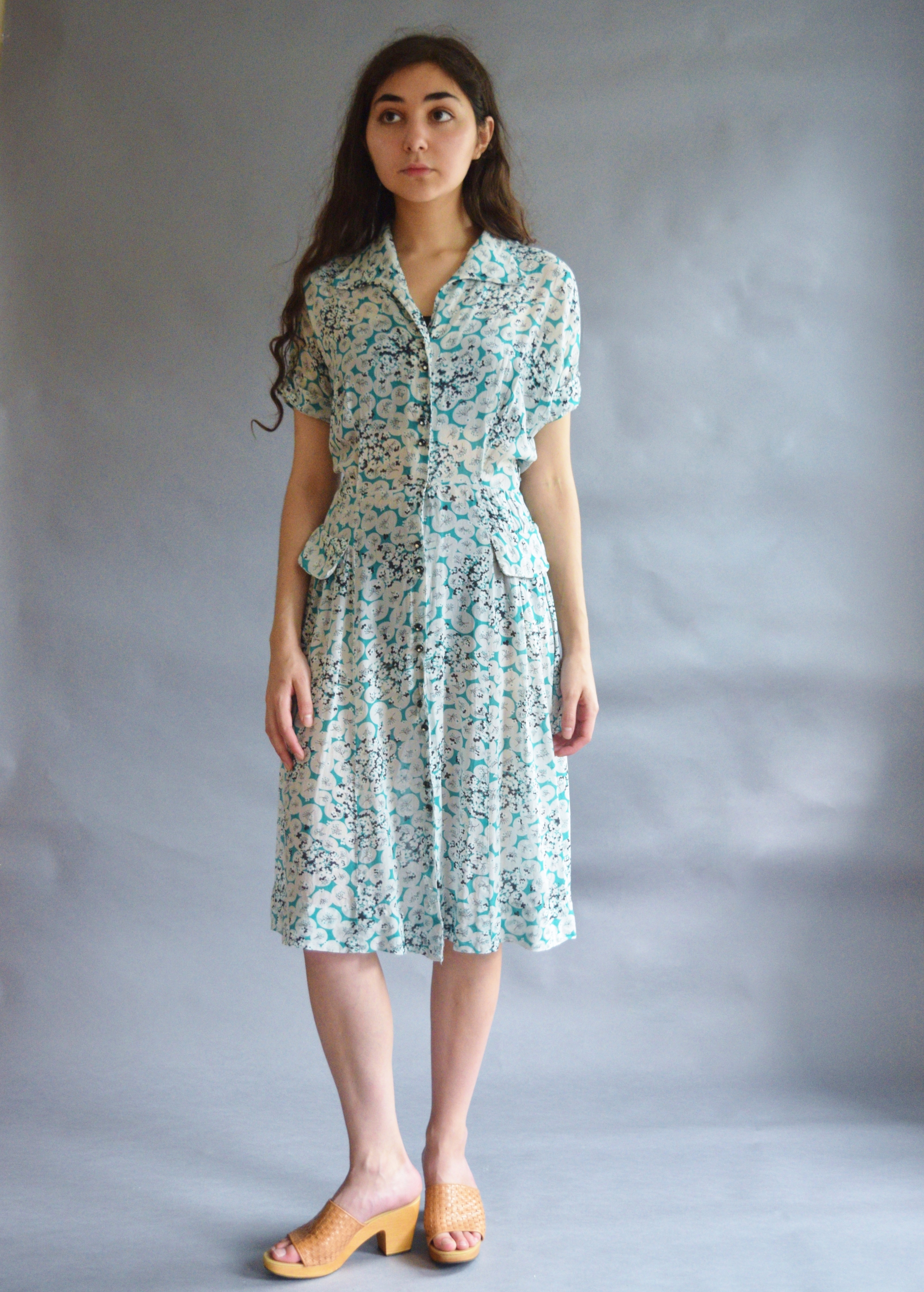 a fulllength studio shot of a woman wearing a vintage 1940sstyle teal and white floral print midi dress with short sleeves and a collared buttondown front. the dress has a pleated skirt and side pockets. she's wearing light brown woven leather clogs or sandals. the background is a plain grey. the model has long dark hair. the overall style is retro or vintage.