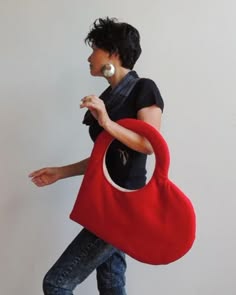 a young woman wearing a black top and blue jeans is holding a large, red heartshaped bag. the bag is made of a soft, plush material and has a white lining. the woman's hair is dark and styled in a short bob. she is looking off to the side, and her expression is relaxed and confident.