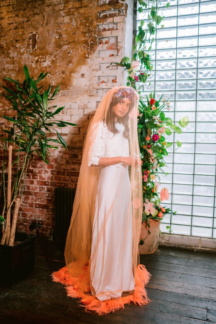 a bride stands in a rustic, industrial setting. she's wearing a flowing white silk wedding gown paired with a long, sheer peachcolored veil that has a vibrant coral feather trim at the bottom.  she also wears a floral crown of colorful blooms.  the background features a brick wall, large plants, and a vibrant floral arrangement against a glassblock window. the overall style is bohemian and vintageinspired. the image is welllit and presents the bride in a stylish and elegant manner.