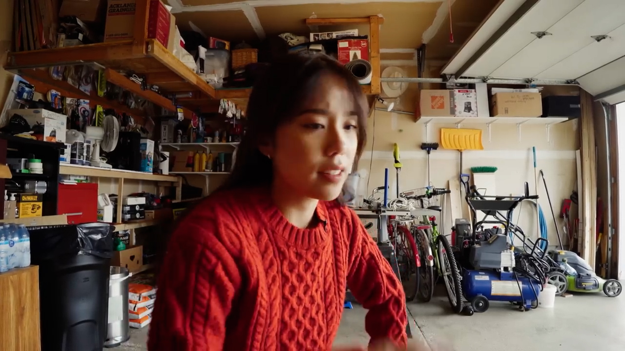 a young asian woman with dark hair in a messy bun is wearing a red cable knit sweater and talking to the camera. it appears to be a selfie or vlog style video. she is in a cluttered garage with a workbench, tools, bikes, and boxes visible in the background. the garage door is open, letting in overcast natural light.  the overall lighting is soft and even.
