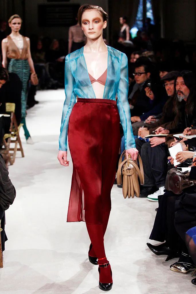 a female model walks the runway in a stunning fashion show. she is wearing a sheer blue top with a red skirt and a pair of black heeled shoes. she is carrying a small handbag in her hand. the model is posing and looking confidently at the camera. the background is blurred, with other models and audience members visible in the distance.  the overall look is elegant and chic, demonstrating a sophisticated and trendy style.