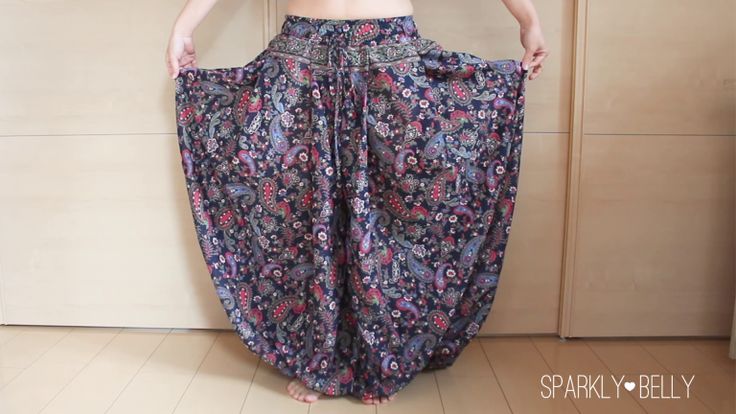 a person stands indoors holding out the sides of a pair of highwaisted harem pants. the pants are made of a dark fabric with a detailed floral and paisley print. the style of the pants is bohemian, featuring a drawstring waist and wide legs. the person is barefoot on a wooden floor.