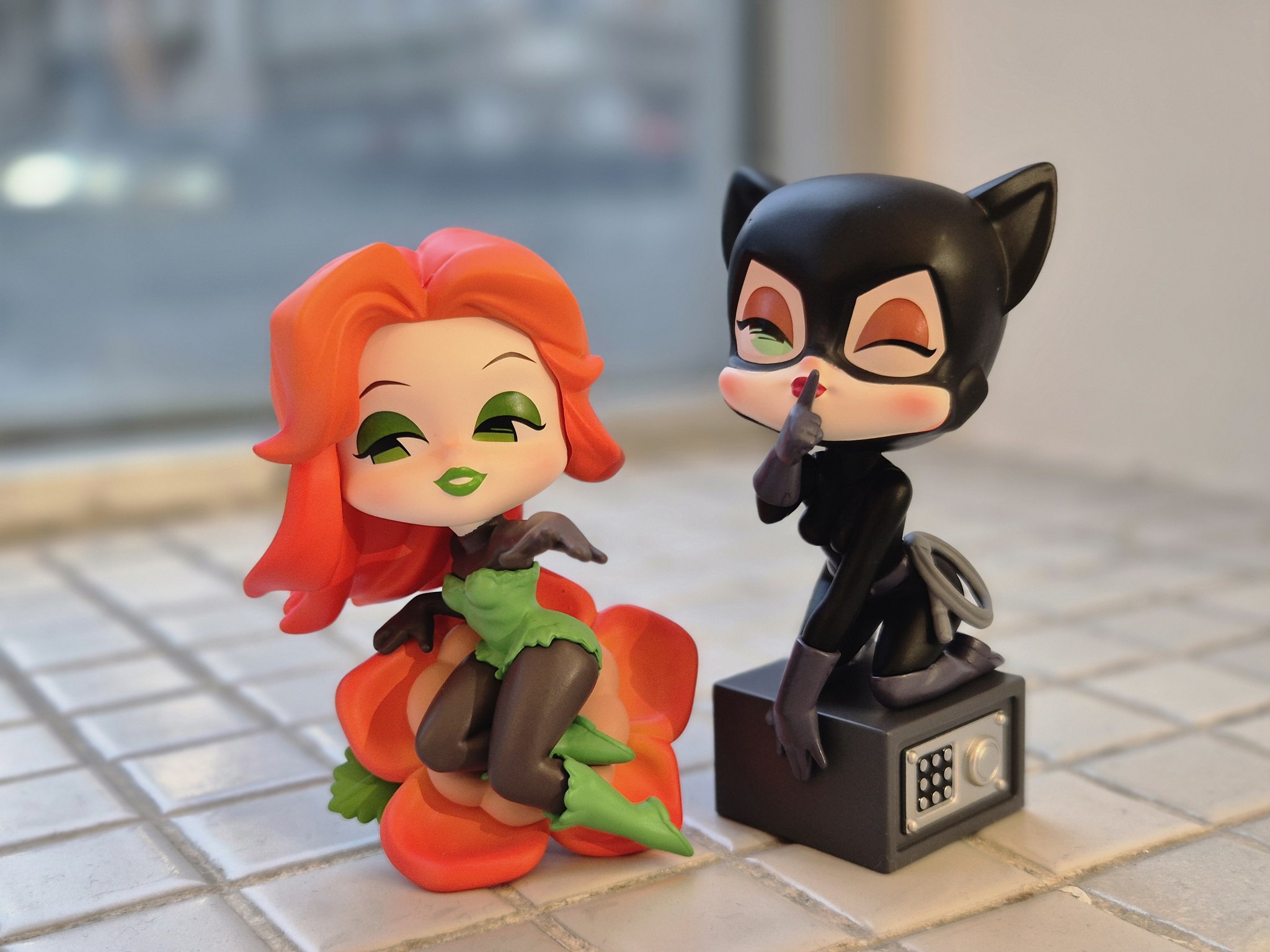 a closeup shot shows two stylized figurines of dc comics villains, poison ivy and catwoman, sitting on a tiled surface. poison ivy, with her distinctive red hair and green accents, is seated on a large orange flower. catwoman, dressed in her classic black suit with cat ears, is posed with a finger to her lips in a shushing gesture, perched atop a small grey safe. the background is blurred, suggesting an indoor setting near a window.