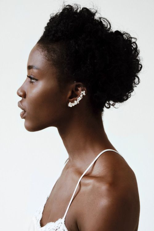 a profile shot of a black woman with dark skin and curly hair styled in an updo. she's wearing a decorative ear cuff, a white top with thin straps, and is set against a white background. the image emphasizes her facial features and the elegance of her style.