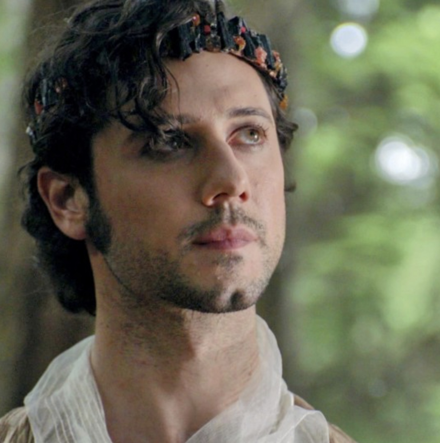 a closeup portrait of a man with dark, curly hair and a crown adorned with dark stones and red accents. he is positioned slightly to the left of the frame, gazing thoughtfully off into the distance with a serene expression. the background is a blur of green foliage, suggesting a forest setting. the soft, natural lighting enhances the contemplative mood of the image.