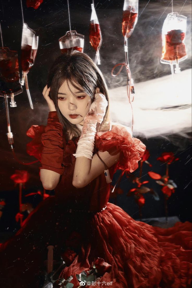 a young woman in a red dress poses dramatically in a dark setting.  she is surrounded by hanging blood bags, and appears to have stage makeup simulating injuries. roses are scattered around her, and the overall atmosphere is dark, gothic, and fantastical, evoking a sense of mystery and perhaps horror.  the image is highly stylized and artistic, with careful attention to lighting and composition.