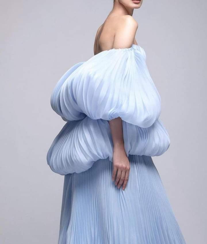 a woman wearing a light blue, offtheshoulder gown with voluminous, pleated sleeves and a pleated skirt. the dress is elegant and appears to be formal wear. the woman is standing against a neutral background.