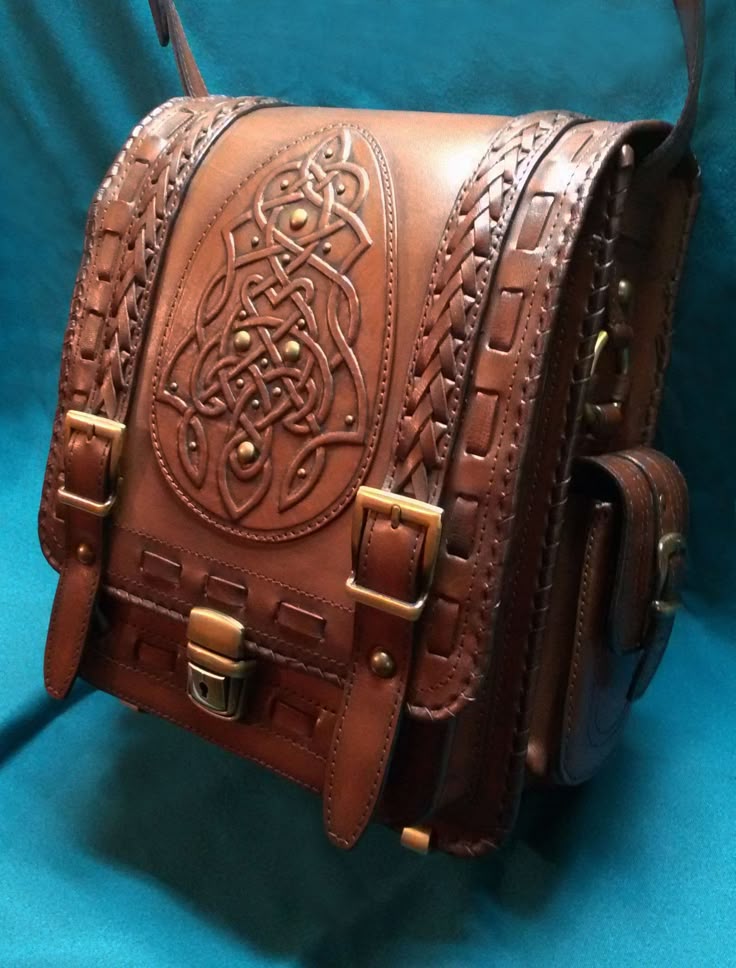 a closeup shot of a brown leather satchel with a celtic knot design embossed on the front flap. the bag has a flap closure with a buckle, two straps with buckles on the front, a shoulder strap, and detailed stitching and braided accents. it is set against a blue background.