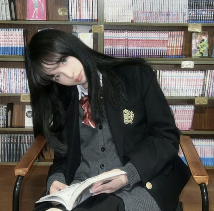 the image shows a young asian girl, possibly a student, sitting in a wooden chair in a library or bookstore. she's wearing a black blazer over a grey cardigan, a typical school uniform style.  her long black hair covers most of her face, partially obscuring her eyes. she is reading a book, and the background is filled with numerous bookshelves stocked with manga or comics. the overall atmosphere is calm and studious.