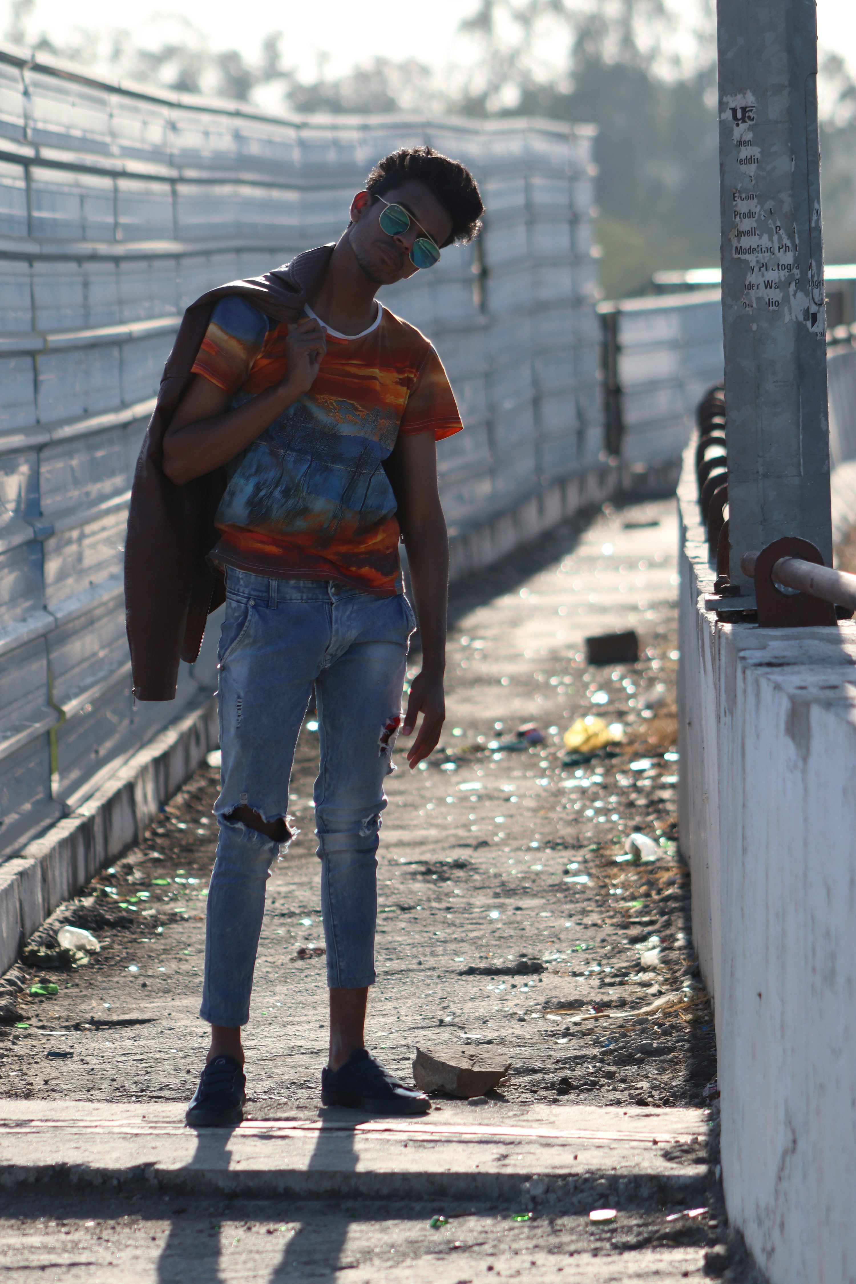 a young man stands on a walkway, casually posing. he's wearing a colorful tshirt, ripped jeans, and a brown leather jacket slung over his shoulder.  he has on sunglasses and darkcolored slipon shoes. the background is an urban setting with a metal fence and concrete walls. the ground is littered with debris and broken glass.  the overall mood is casual and stylish.